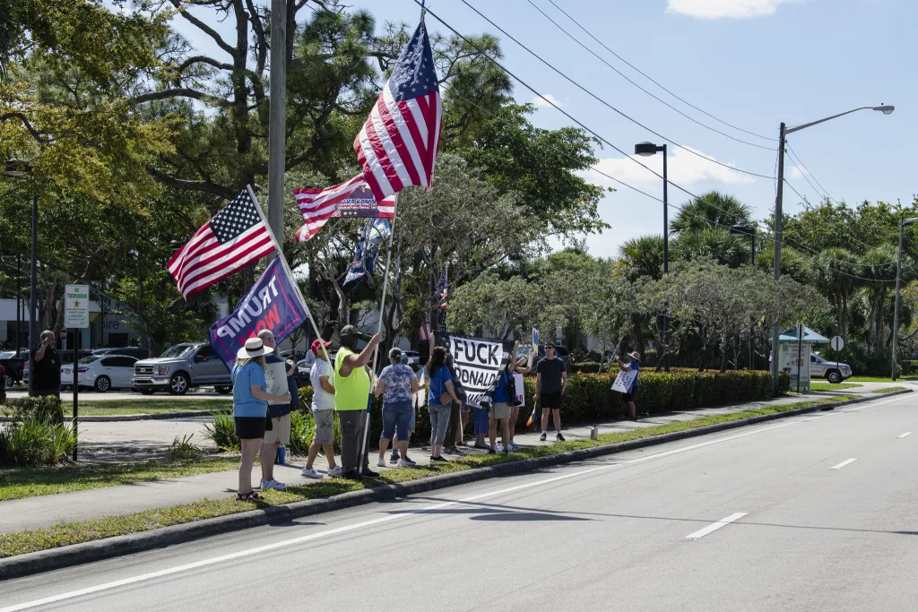 Protesters and MAGA supporters face off outside Trump’s golf course, waving American flags and holding signs with strong political statements. A large "F*** Donald Trump" banner stands in contrast to Trump 2024 flags, highlighting the deep political divide.