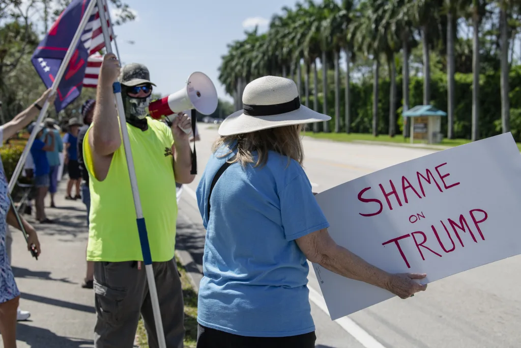 A protester outside Trump International Golf Club in West Palm Beach, FL, on March 2, 2025, holds a sign reading "Shame on Trump" while a counter-protester in a neon shirt and skull-patterned face covering uses a megaphone. Behind them, other demonstrators wave Trump flags, underscoring the tension between opposing groups.