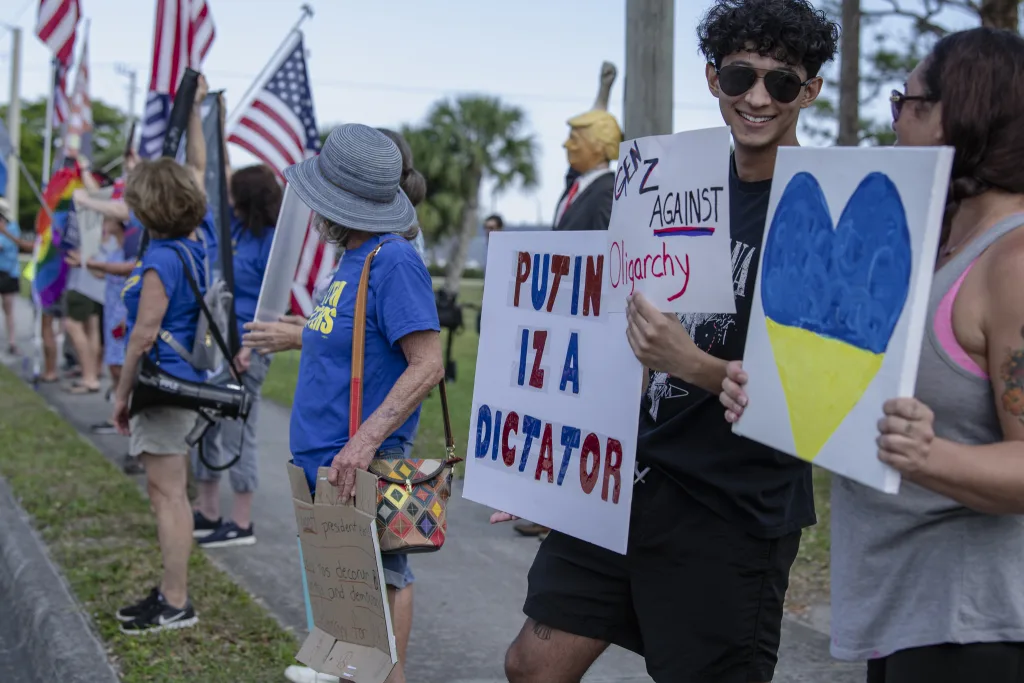  A group of protesters, including young activists, holds signs supporting Ukraine and democracy, with bold messages like “Putin is a dictator.” In the background, a strange effigy of Trump tied to a pole stands among MAGA counterprotesters, symbolizing the tense standoff between the two groups.