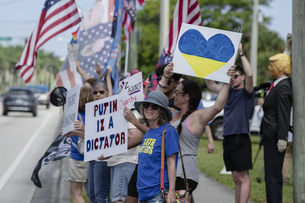 Protesters send the president a message outside Trump International Golf Course, holding signs for democracy and Ukraine, while MAGA counter-protesters wave flags in opposition. A striking image in the background shows a Trump effigy tied to a pole, adding another layer to the confrontation.