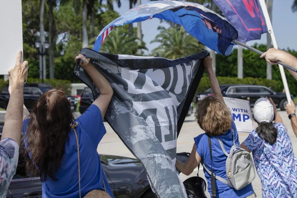 Counter-protesters outside Trump International Golf Club in West Palm Beach, FL, on March 2, 2025, use flags to block a protester’s sign from Trump's view as he drives by. The scene captures the tension between activists standing against Trump and those attempting to silence them.  Join the Protest for Justice