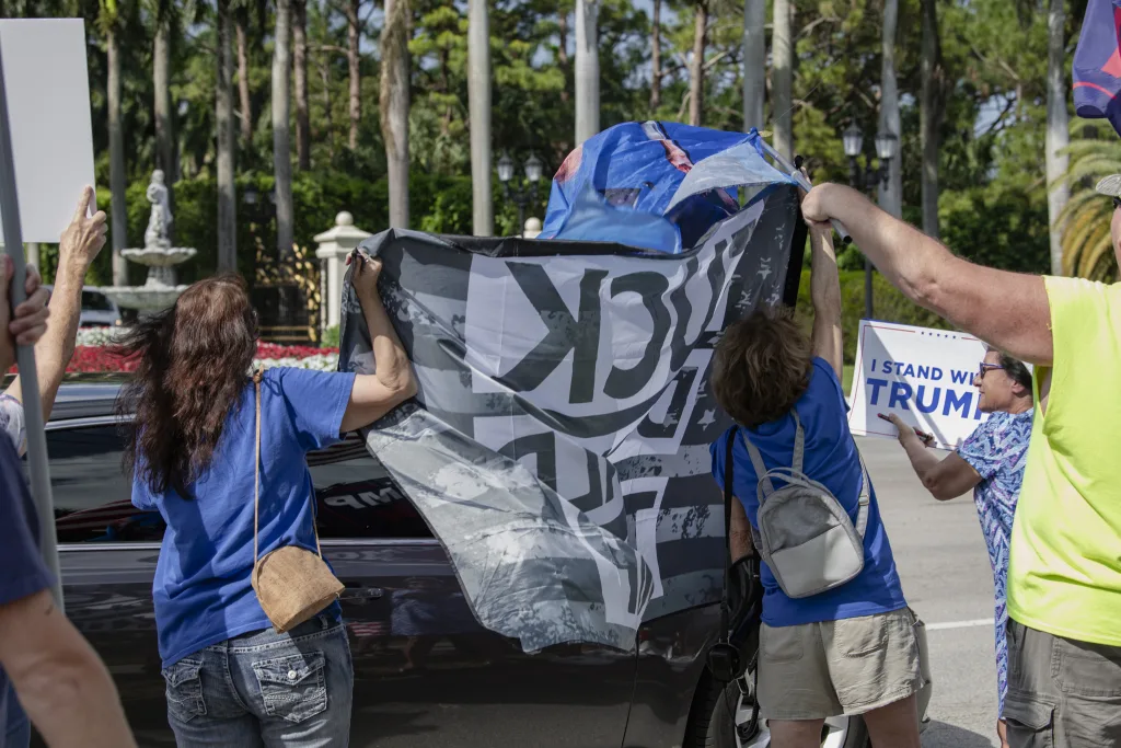 Protesters outside Trump International Golf Club in West Palm Beach, FL, confront counter-protesters attempting to obstruct their message. A group of activists in blue push back against Trump supporters waving flags to cover their signs. The scene captures the ongoing struggle for democracy and free speech.